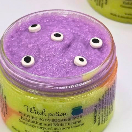 A whipped sugar scrub in a clear jar with layered light green, orange, purple, and yellow colors, topped with an eyeball candy decoration.