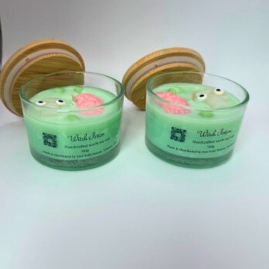 A light green, single-wick candle in a clear jar with a bamboo lid, featuring a skull, pink brain, and bubbles decoration on top—perfect for Halloween.