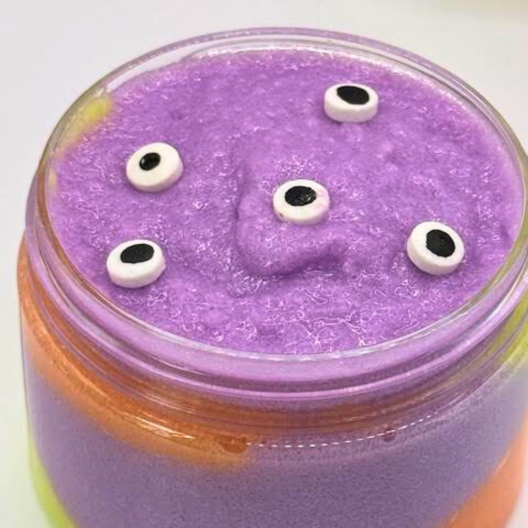 A whipped sugar scrub in a clear jar with layered light green, orange, purple, and yellow colors, topped with an eyeball candy decoration.