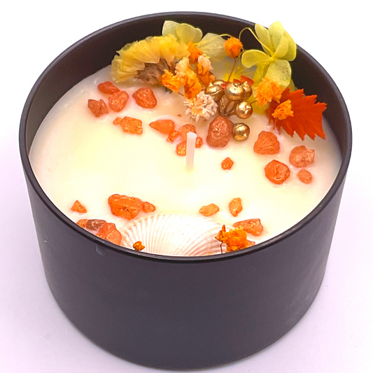 A single-wick white candle in a black jar, topped with dried yellow and orange florals, leaves, golden rocks, and a seashell decoration.