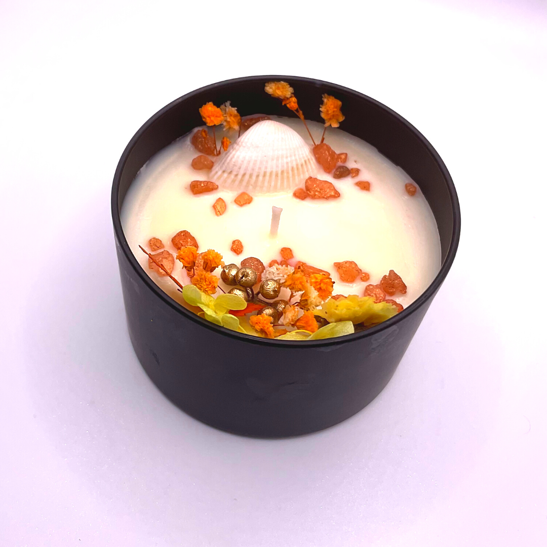 A single-wick white candle in a black jar, topped with dried yellow and orange florals, leaves, golden rocks, and a seashell decoration.