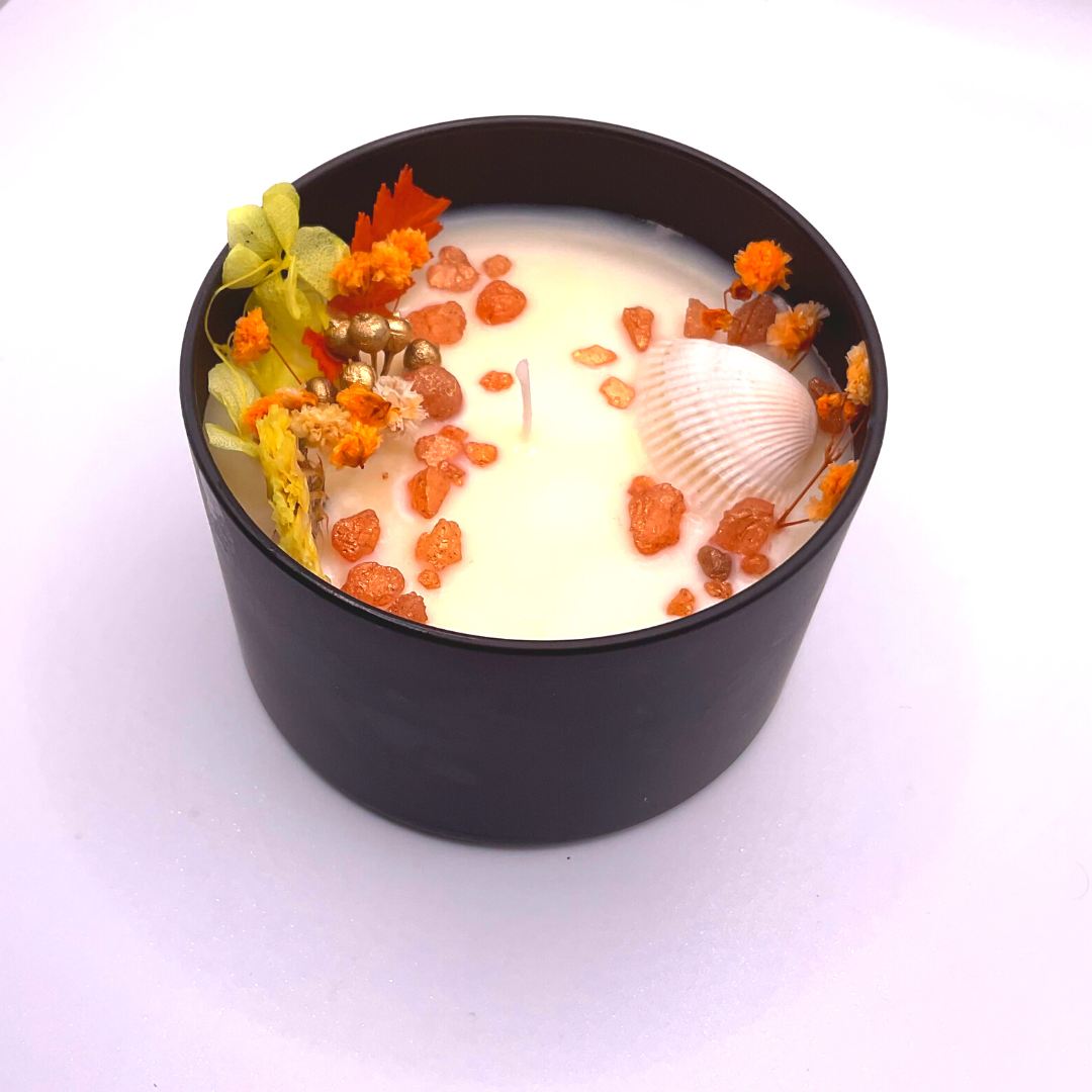 A single-wick white candle in a black jar, topped with dried yellow and orange florals, leaves, golden rocks, and a seashell decoration.