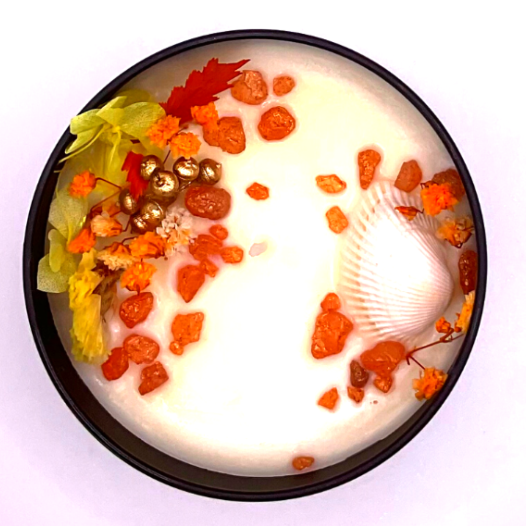 A single-wick white candle in a black jar, topped with dried yellow and orange florals, leaves, golden rocks, and a seashell decoration.
