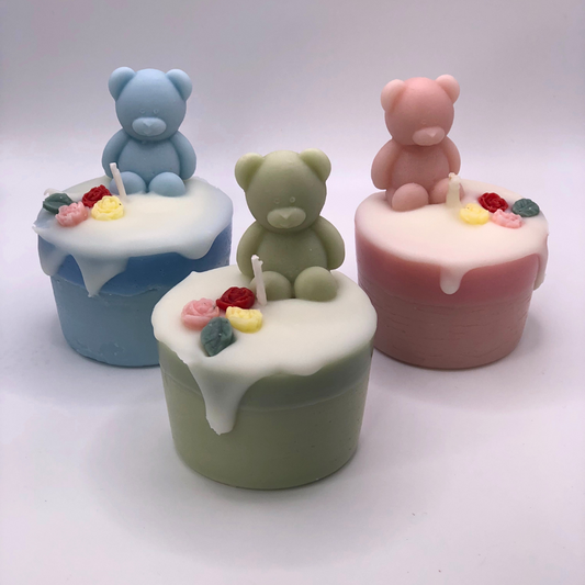 A set of three cupcake candles in pink, light green, and baby blue, each decorated with a teddy bear and flowers on top.