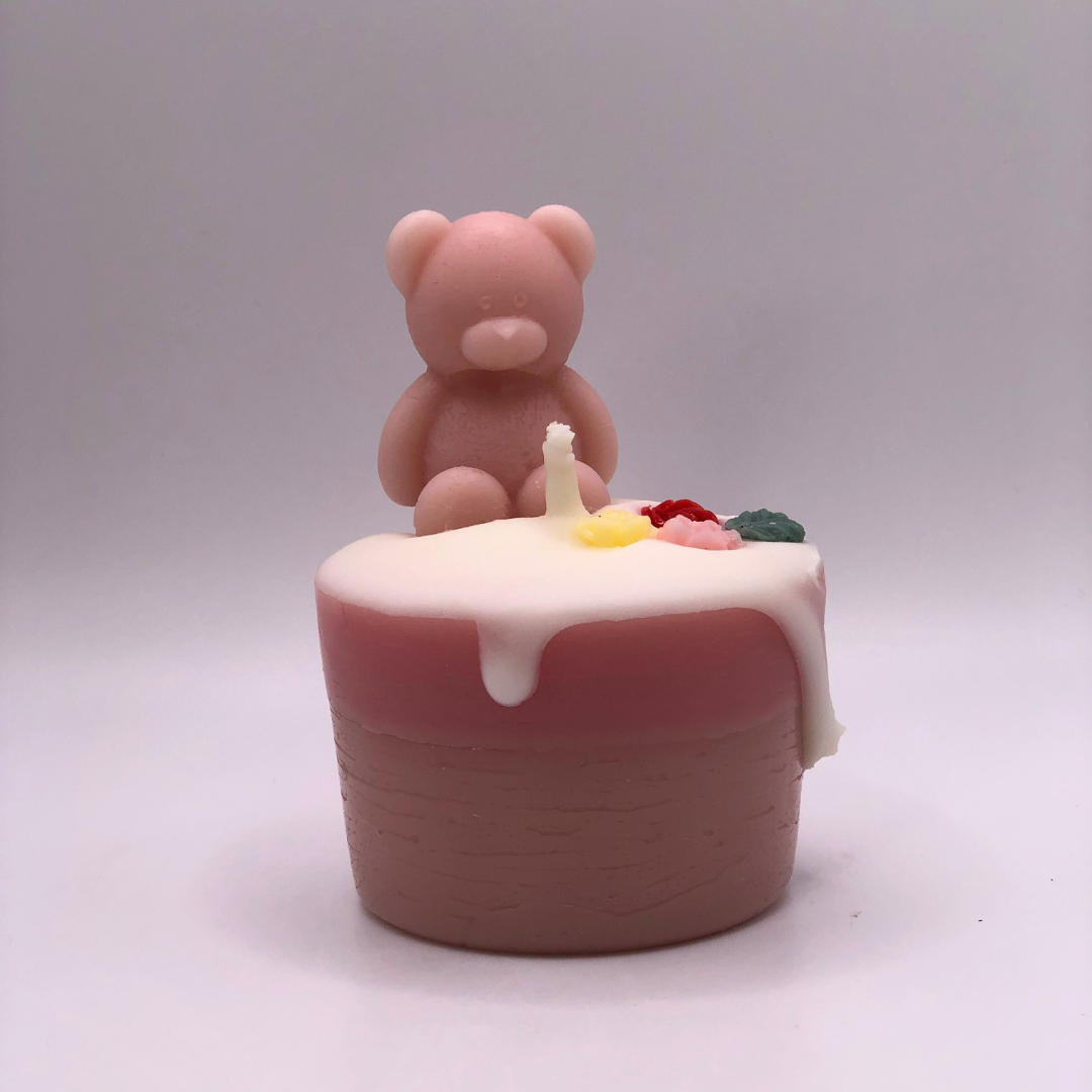 A single wick cupcake candle in pink, decorated with a teddy bear and flowers on top.
