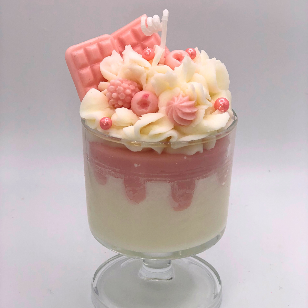 A single-wick candle in a dessert glass with a white bottom, pink middle layer, and topped with white whipped icing wax, decorated with Chocolate bar-shaped, raspberry-shaped, blueberry-shaped, and meringue candy-shaped candles, and pearl candy