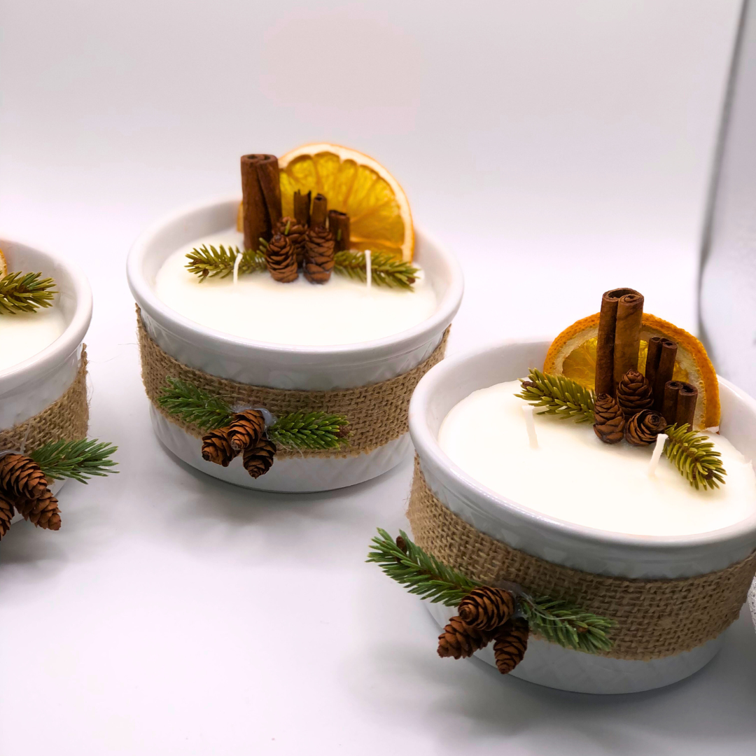 A triple-wick white candle in a white container, decorated with a ribbon, dry orange wedge, cinnamon sticks, pinecones, and pine needle branches.