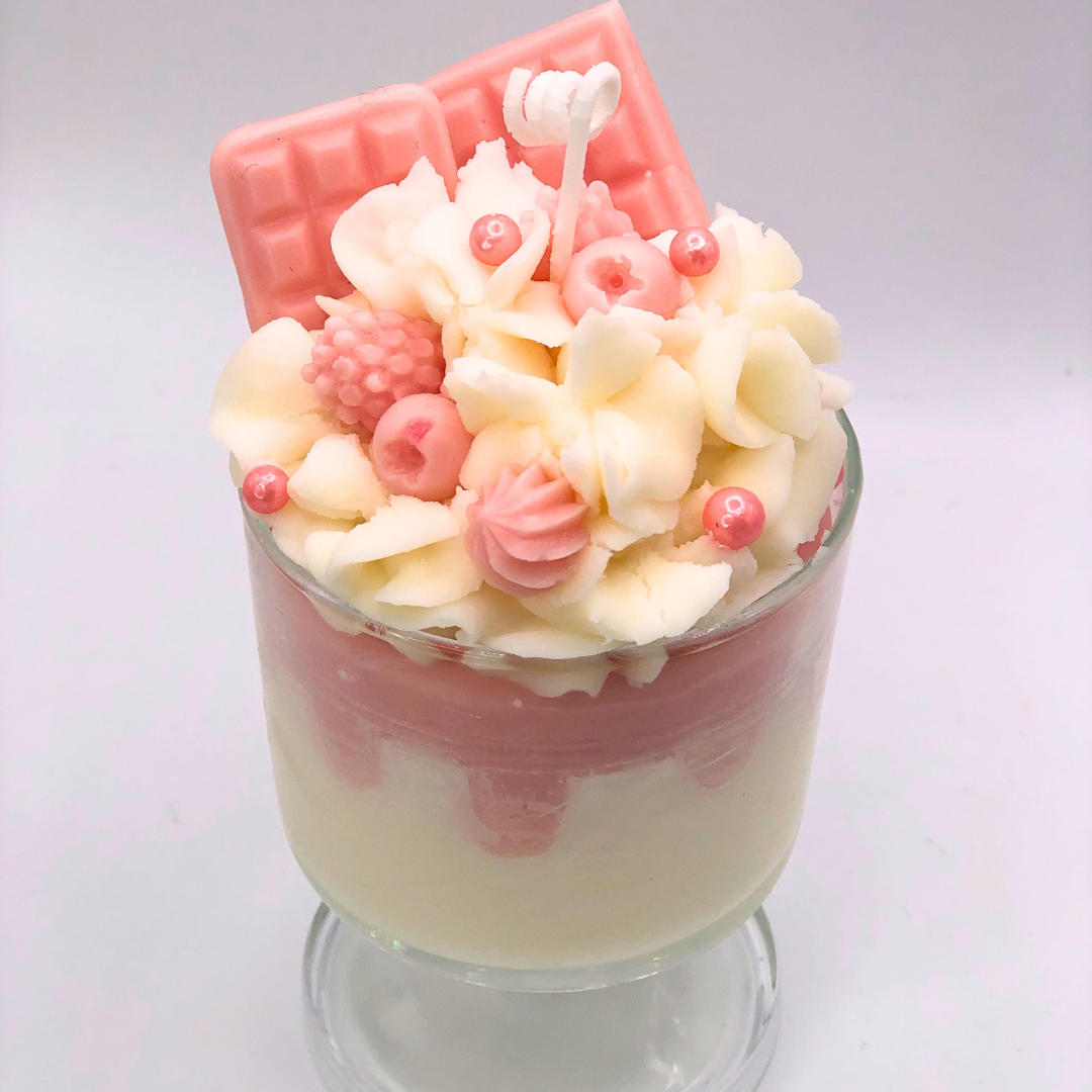 A single-wick candle in a dessert glass with a white bottom, pink middle layer, and topped with white whipped icing wax, decorated with Chocolate bar-shaped, raspberry-shaped, blueberry-shaped, and meringue candy-shaped candles, and pearl candy