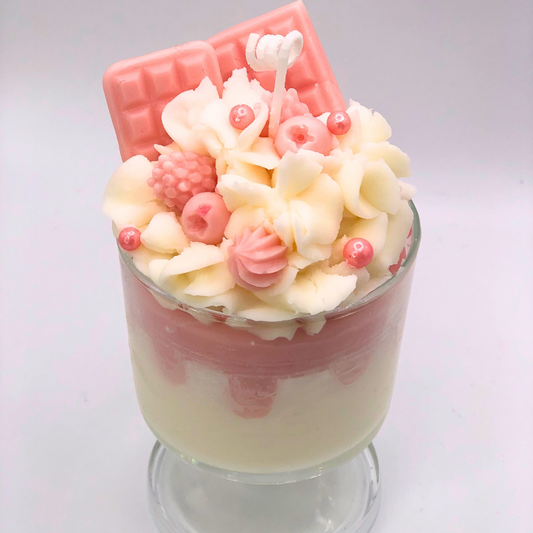 A single-wick candle in a dessert glass with a white bottom, pink middle layer, and topped with white whipped icing wax, decorated with Chocolate bar-shaped, raspberry-shaped, blueberry-shaped, and meringue candy-shaped candles, and pearl candy