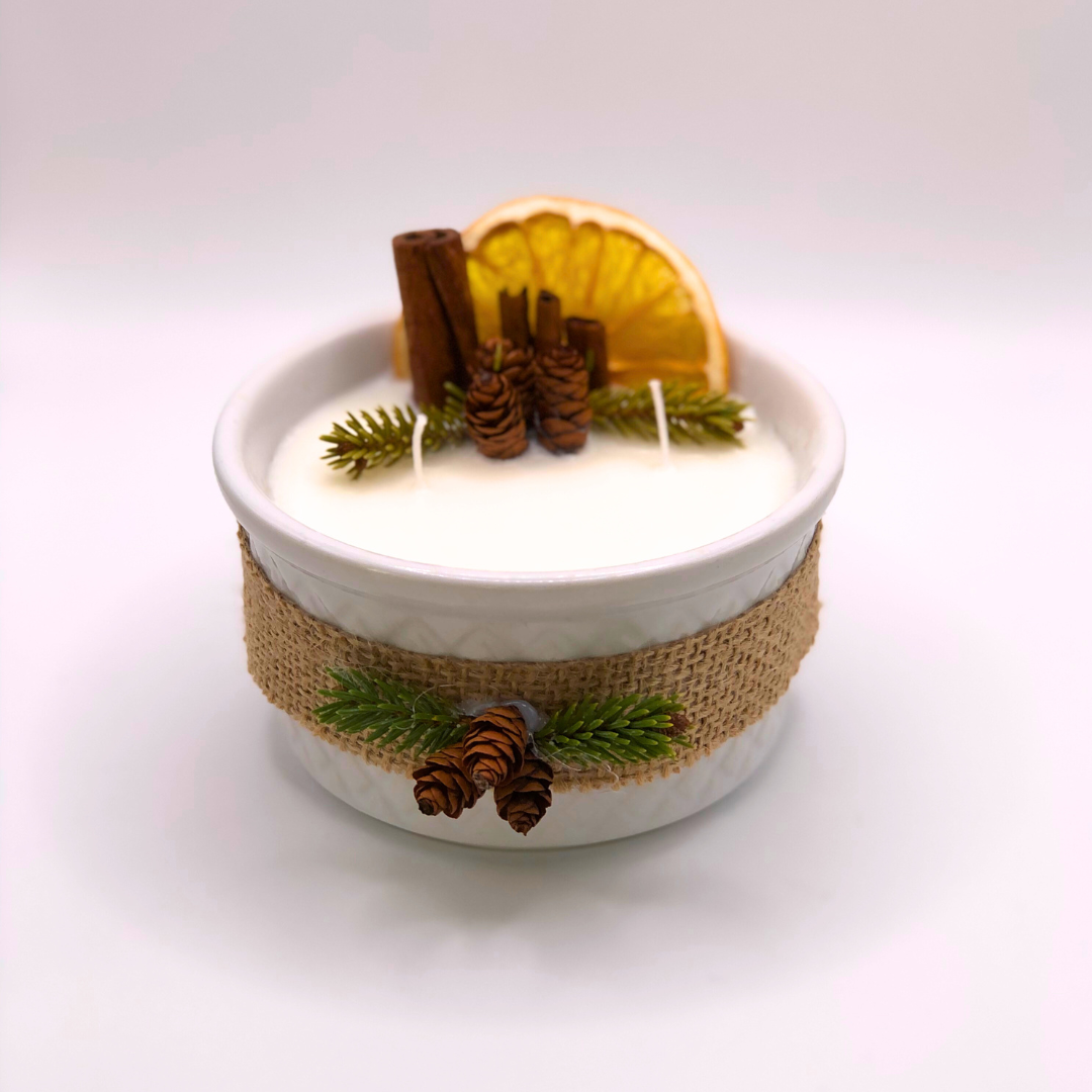 A triple-wick white candle in a white container, decorated with a ribbon, dry orange wedge, cinnamon sticks, pinecones, and pine needle branches.