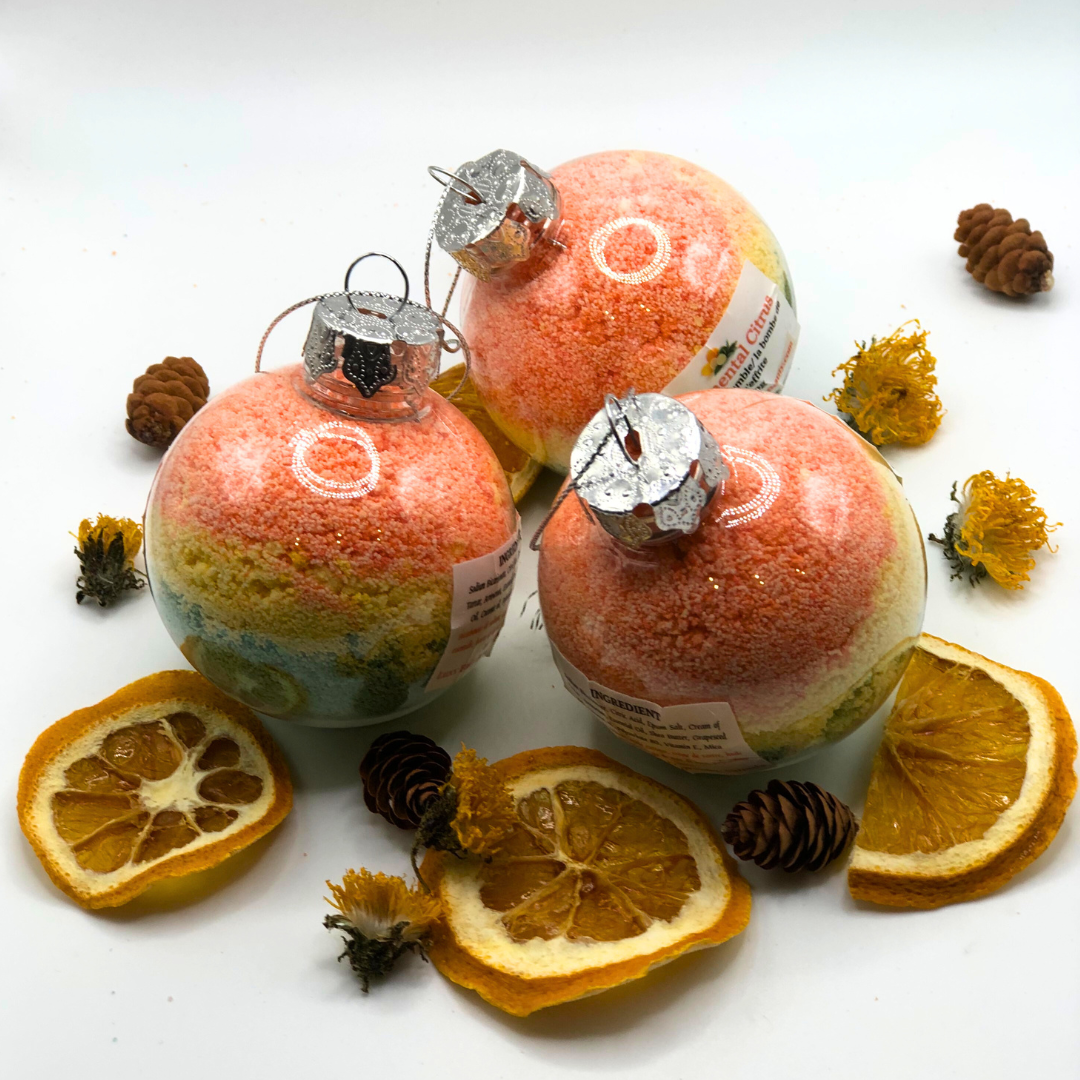 A bath crumble in yellow, green, and orange colors placed inside a Christmas tree ornament, decorated with a pinecone, dried yellow flowers, and dried orange slices.