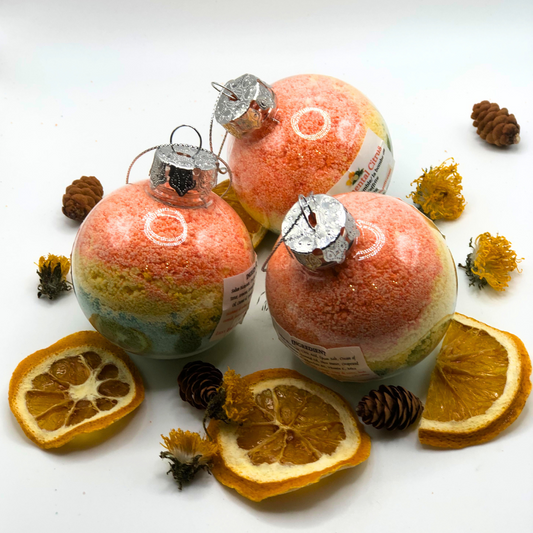 A bath crumble in yellow, green, and orange colors placed inside a Christmas tree ornament, decorated with a pinecone, dried yellow flowers, and dried orange slices.