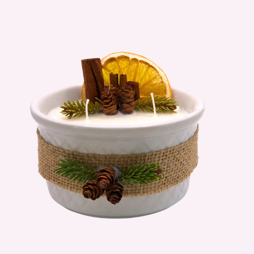 A triple-wick white candle in a white container, decorated with a ribbon, dry orange wedge, cinnamon sticks, pinecones, and pine needle branches.