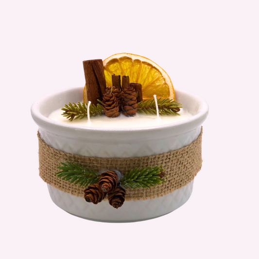 A triple-wick white candle in a white container, decorated with a ribbon, dry orange wedge, cinnamon sticks, pinecones, and pine needle branches.