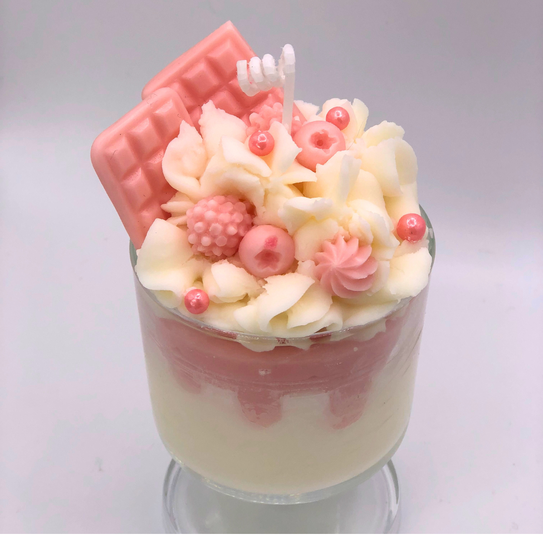 A single-wick candle in a dessert glass with a white bottom, pink middle layer, and topped with white whipped icing wax, decorated with Chocolate bar-shaped, raspberry-shaped, blueberry-shaped, and meringue candy-shaped candles, and pearl candy