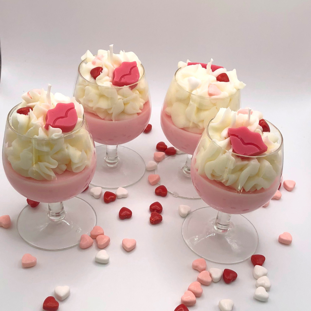 A single-wick candle in a wine glass with a pink bottom, topped with whipped wax icing, decorated with a red lip-shaped candle and heart-shaped candy.