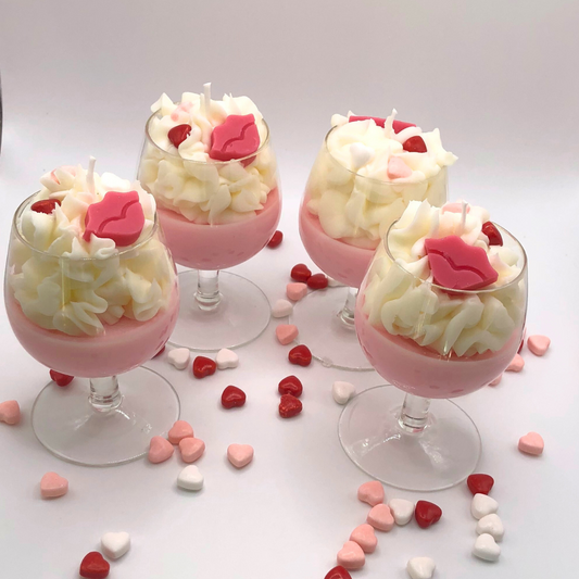 A single-wick candle in a wine glass with a pink bottom, topped with whipped wax icing, decorated with a red lip-shaped candle and heart-shaped candy.