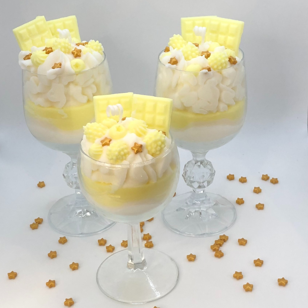 A single-wick candle in a dessert glass with a white bottom, and yellow middle layer, and topped with white whipped icing wax, decorated with Yellow Chocolate bar-shaped, raspberry-shaped, blueberry-shaped, and meringue candy-shaped candles, and pearl star candy