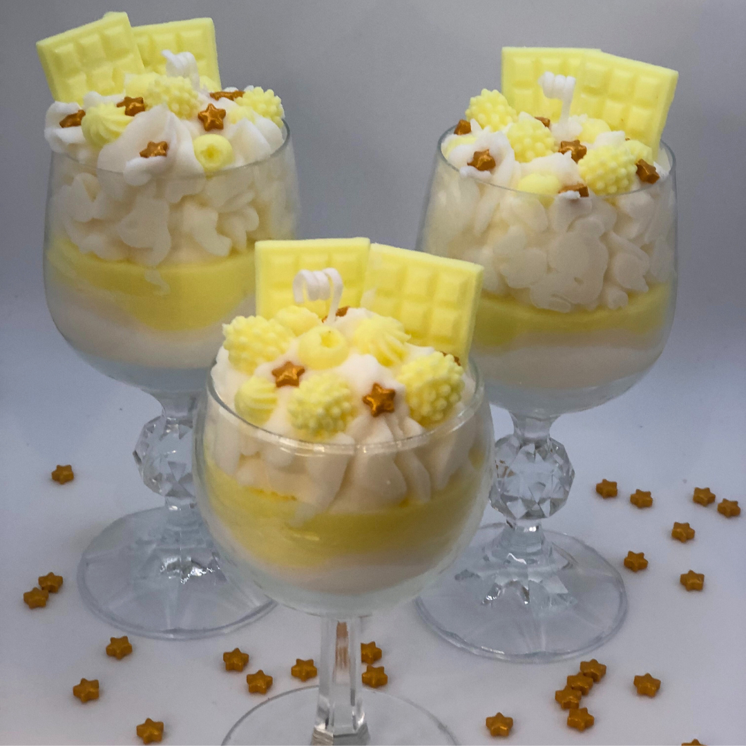 A single-wick candle in a dessert glass with a white bottom, and yellow middle layer, and topped with white whipped icing wax, decorated with Yellow Chocolate bar-shaped, raspberry-shaped, blueberry-shaped, and meringue candy-shaped candles, and pearl star candy