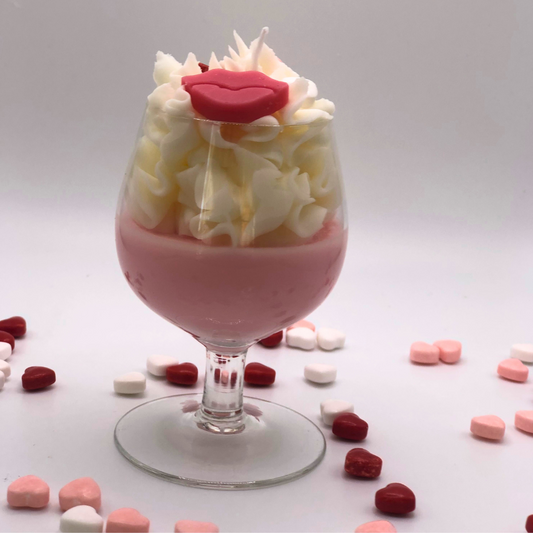 A single-wick candle in a wine glass with a pink bottom, topped with whipped wax icing, decorated with a red lip-shaped candle and heart-shaped candy.