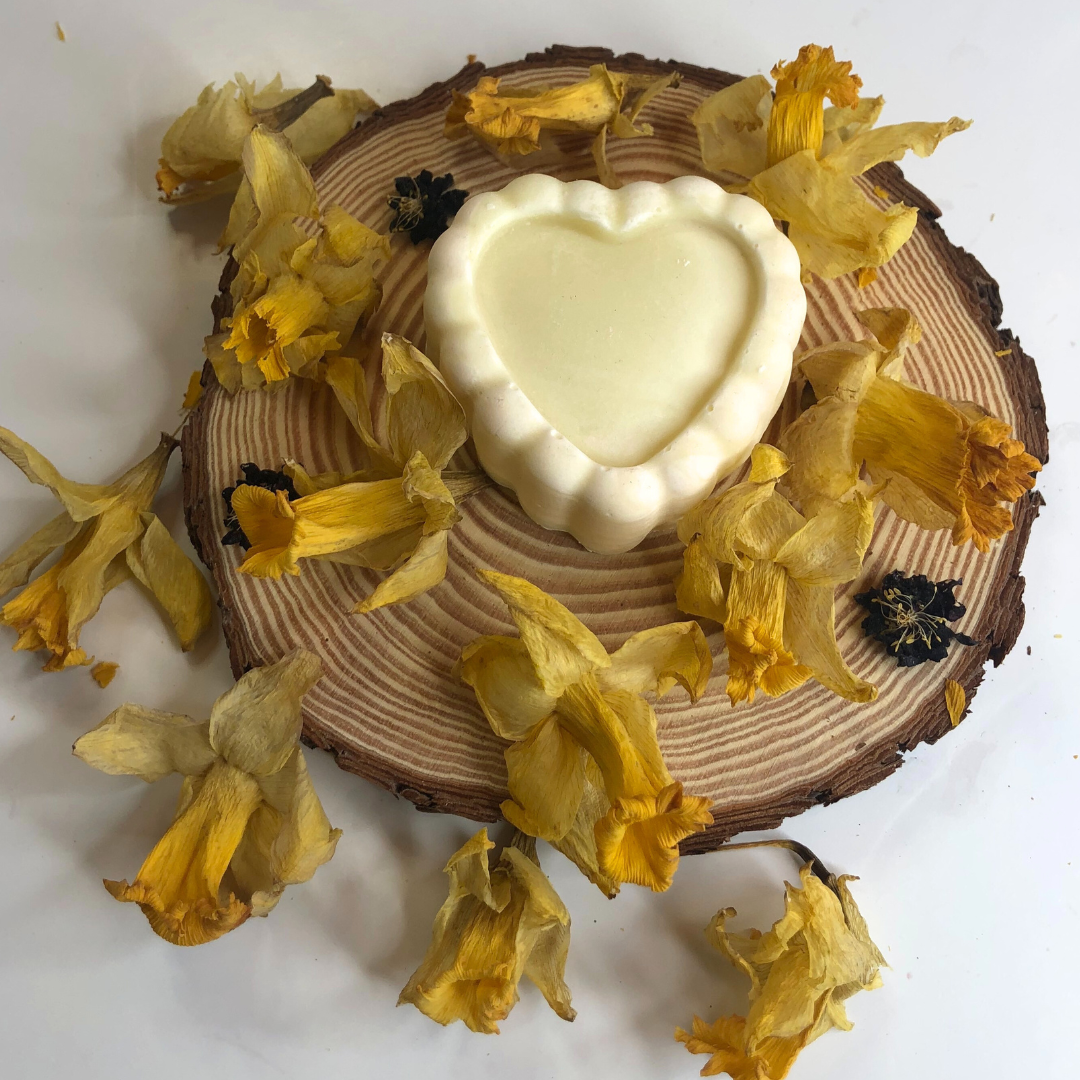 A white heart-shaped soap bar placed on a piece of wood, decorated with dried yellow flowers.