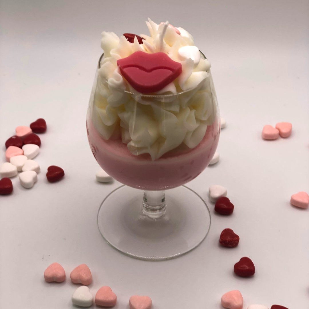 A single-wick candle in a wine glass with a pink bottom, topped with whipped wax icing, decorated with a red lip-shaped candle and heart-shaped candy.