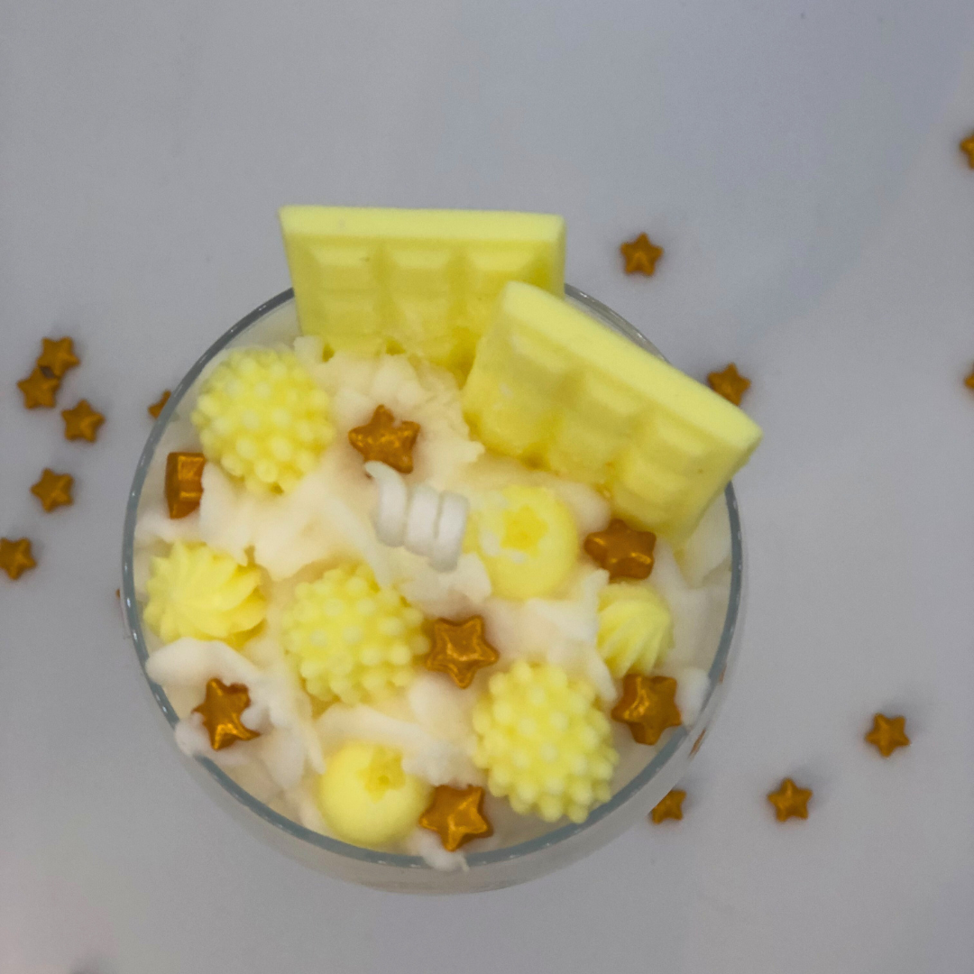 A single-wick candle in a dessert glass with a white bottom, and yellow middle layer, and topped with white whipped icing wax, decorated with Yellow Chocolate bar-shaped, raspberry-shaped, blueberry-shaped, and meringue candy-shaped candles, and pearl star candy