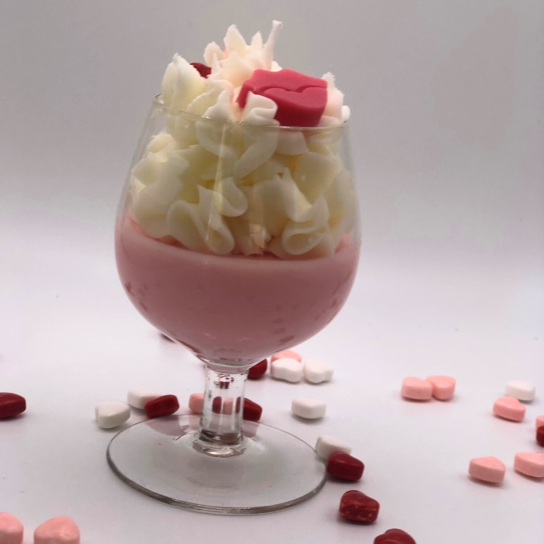 A single-wick candle in a wine glass with a pink bottom, topped with whipped wax icing, decorated with a red lip-shaped candle and heart-shaped candy.