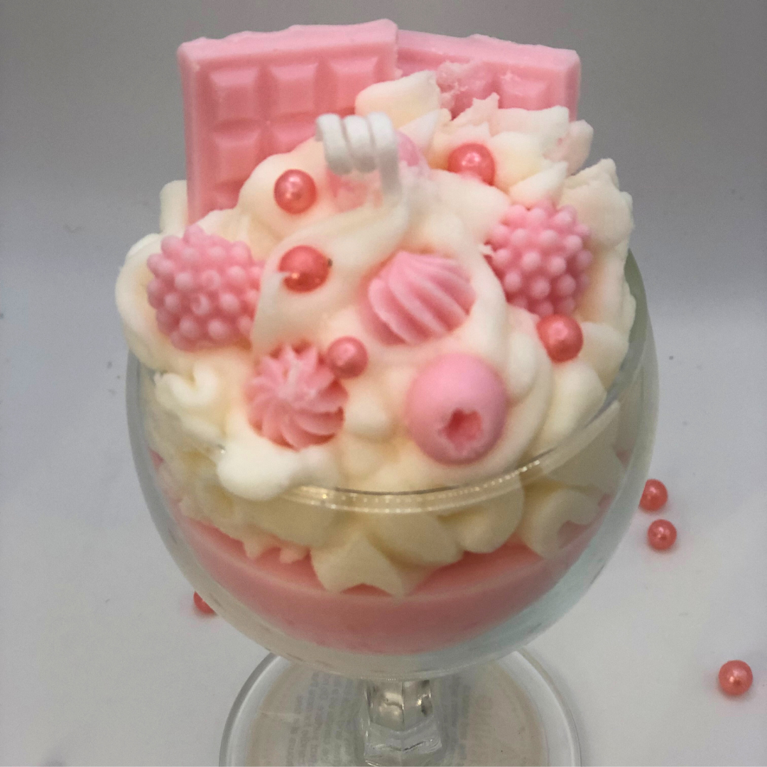 A single-wick candle in a dessert glass with a white bottom, pink middle layer, and topped with white whipped icing wax, decorated with Chocolate bar-shaped, raspberry-shaped, blueberry-shaped, and meringue candy-shaped candles, and pearl candy