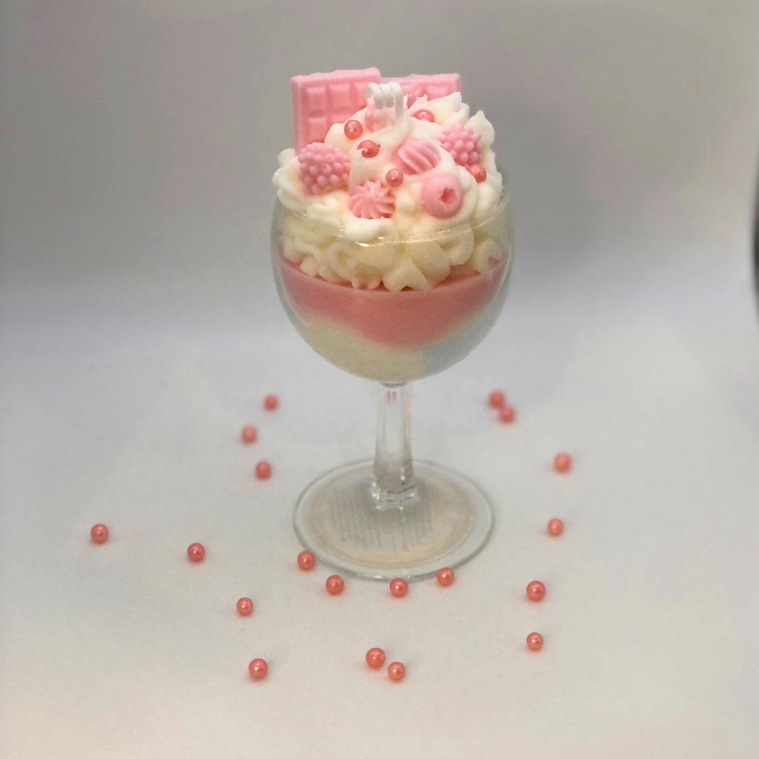 A single-wick candle in a dessert glass with a white bottom, pink middle layer, and topped with white whipped icing wax, decorated with Chocolate bar-shaped, raspberry-shaped, blueberry-shaped, and meringue candy-shaped candles, and pearl candy