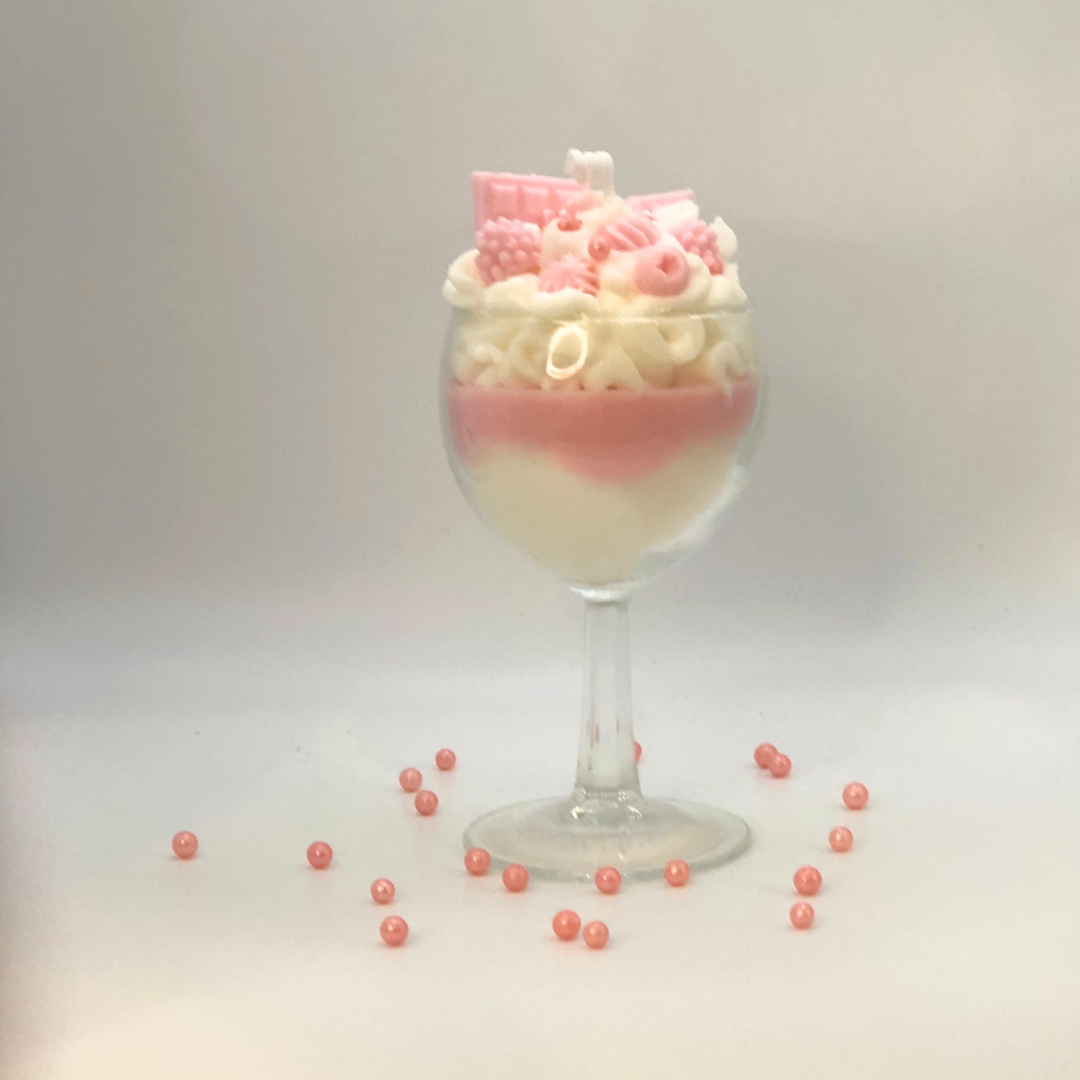 A single-wick candle in a dessert glass with a white bottom, pink middle layer, and topped with white whipped icing wax, decorated with Chocolate bar-shaped, raspberry-shaped, blueberry-shaped, and meringue candy-shaped candles, and pearl candy