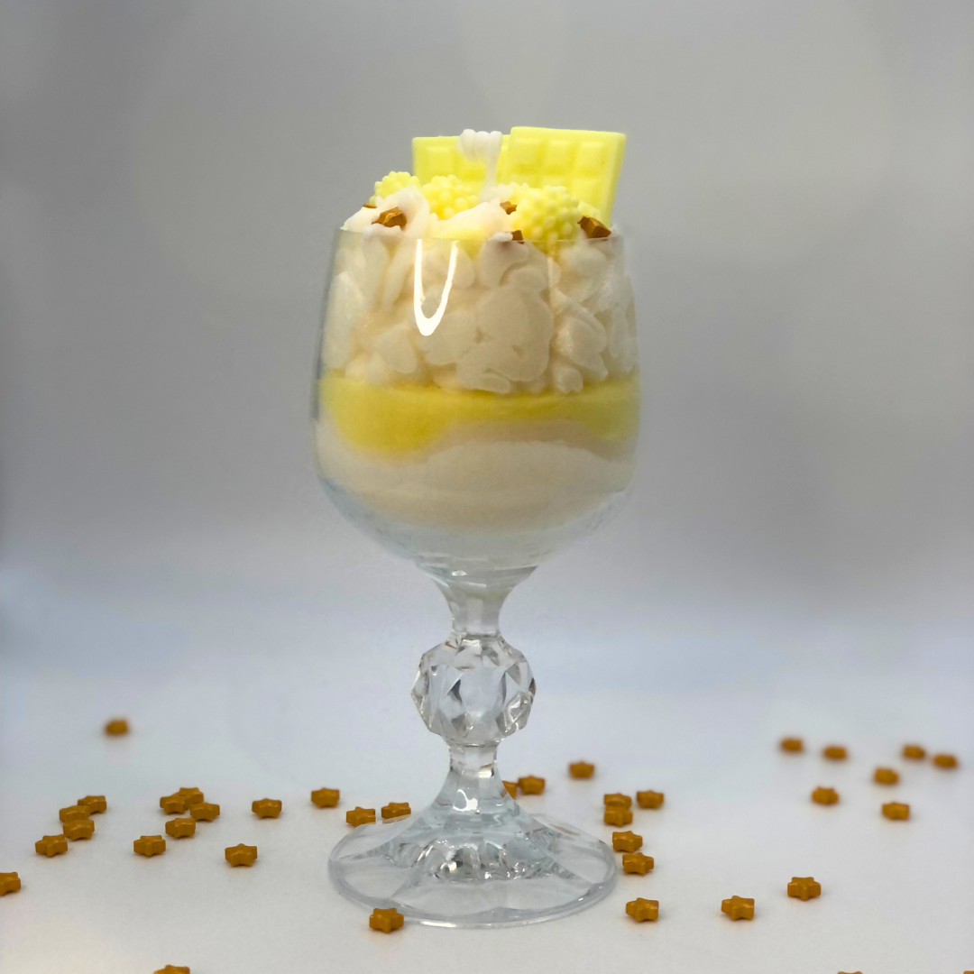 A single-wick candle in a dessert glass with a white bottom, and yellow middle layer, and topped with white whipped icing wax, decorated with Yellow Chocolate bar-shaped, raspberry-shaped, blueberry-shaped, and meringue candy-shaped candles, and pearl star candy