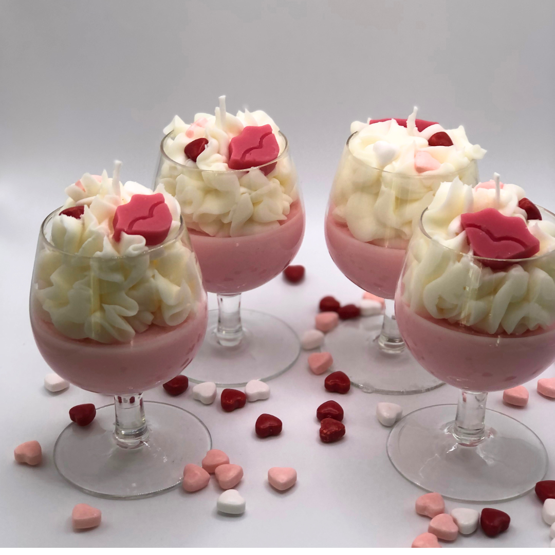 A single-wick candle in a wine glass with a pink bottom, topped with whipped wax icing, decorated with a red lip-shaped candle and heart-shaped candy.
