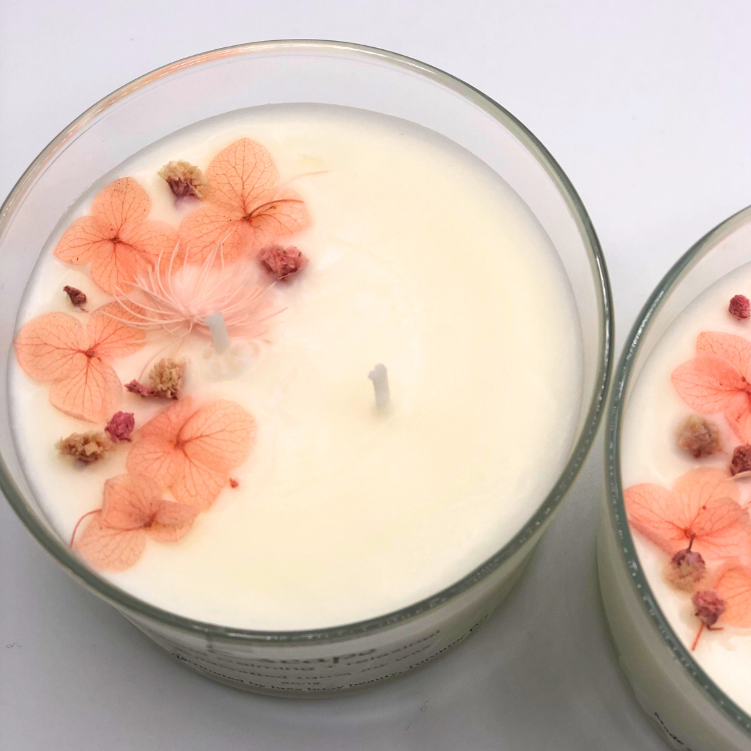 A single-wick white candle in a clear jar, topped with a dried pink floral decoration.