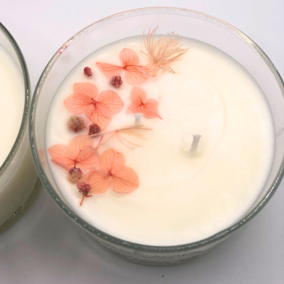 A single-wick white candle in a clear jar, topped with a dried pink floral decoration.