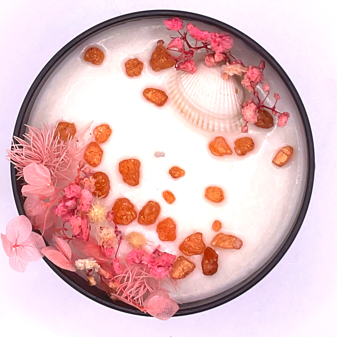 A double-wick white candle in a black jar, topped with dried pink florals, small seashells, and golden rocks.