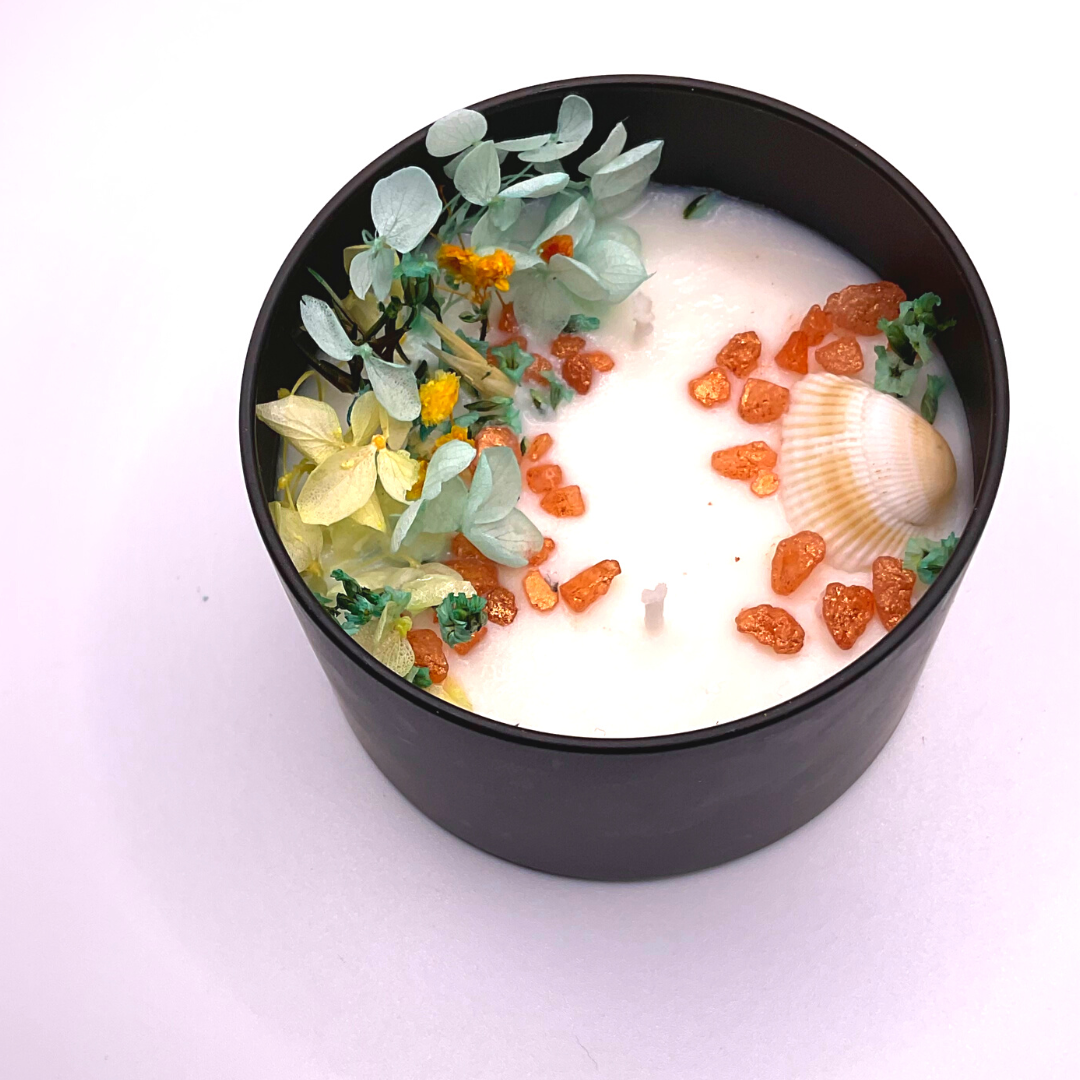 A double-wick white candle in a black jar, topped with dried yellow and blue florals, small seashells, and golden rocks.