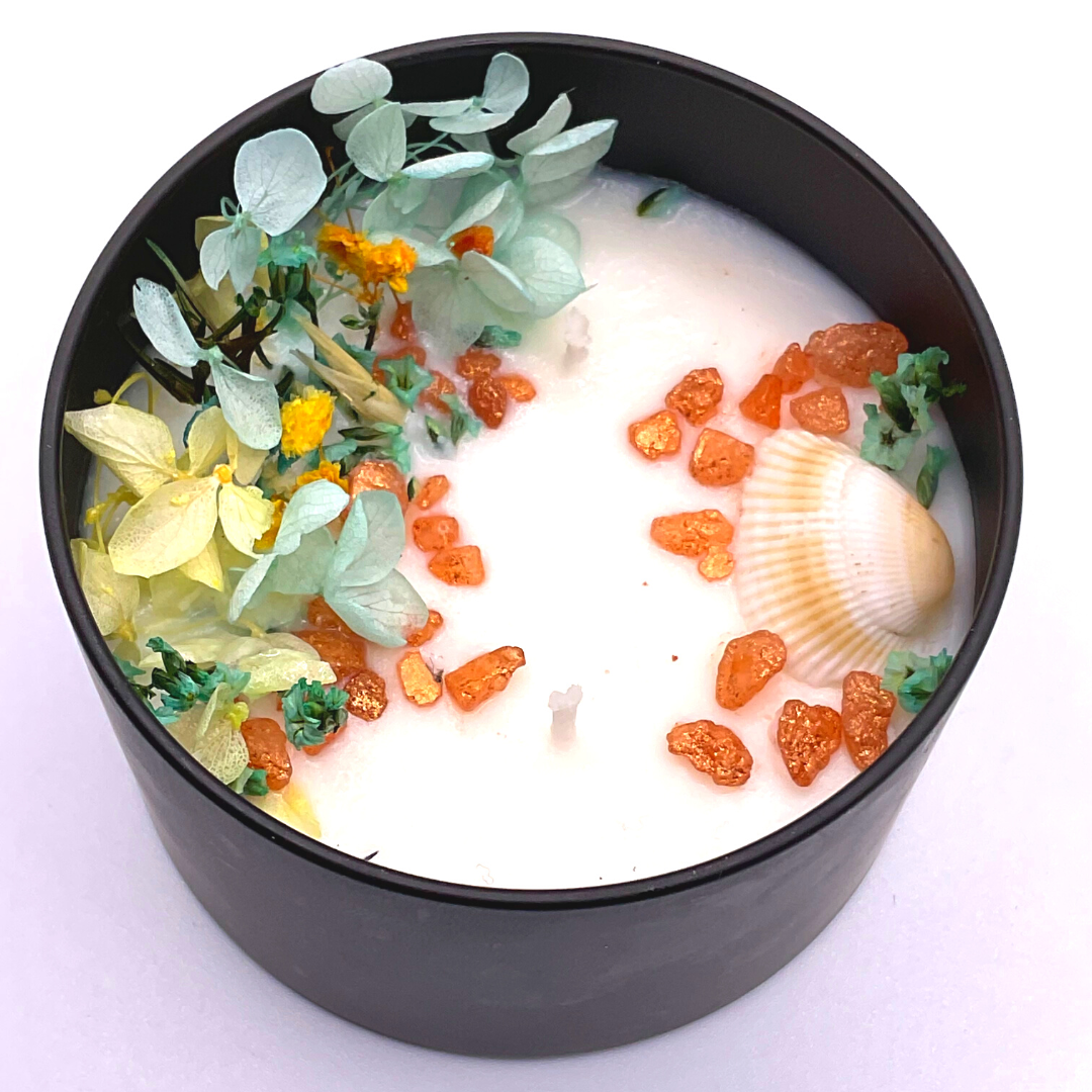A double-wick white candle in a black jar, topped with dried yellow and blue florals, small seashells, and golden rocks.