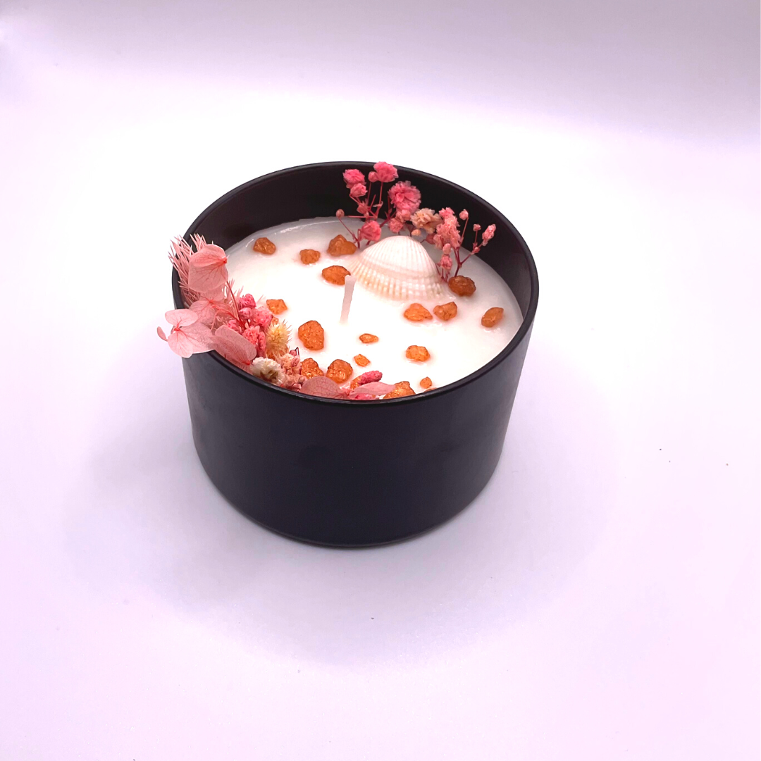 A double-wick white candle in a black jar, topped with dried pink florals, small seashells, and golden rocks.
