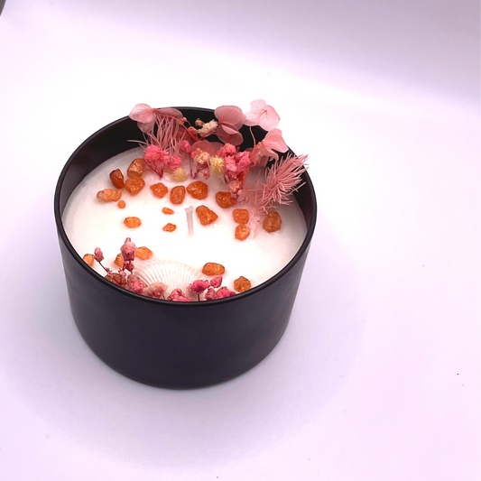 A double-wick white candle in a black jar, topped with dried pink florals, small seashells, and golden rocks.
