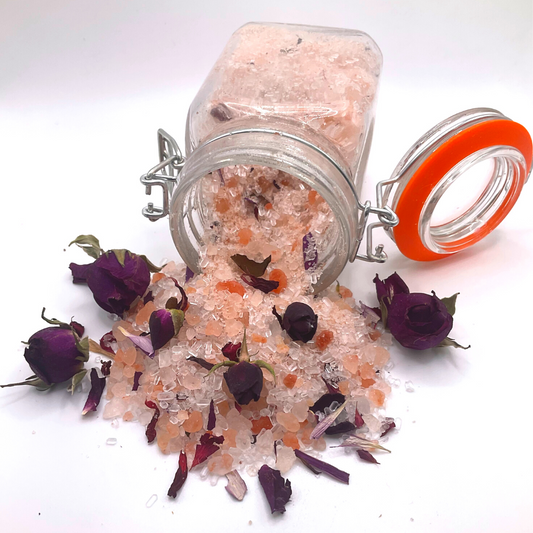 A flipped-over clear jar spilling pink Himalayan salts mixed with dried florals and rose buds.
