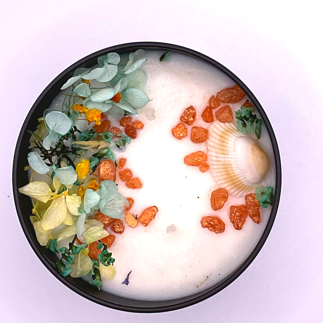 A double-wick white candle in a black jar, topped with dried yellow and blue florals, small seashells, and golden rocks.