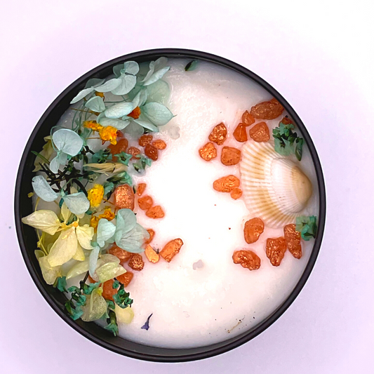 A double-wick white candle in a black jar, topped with dried yellow and blue florals, small seashells, and golden rocks.