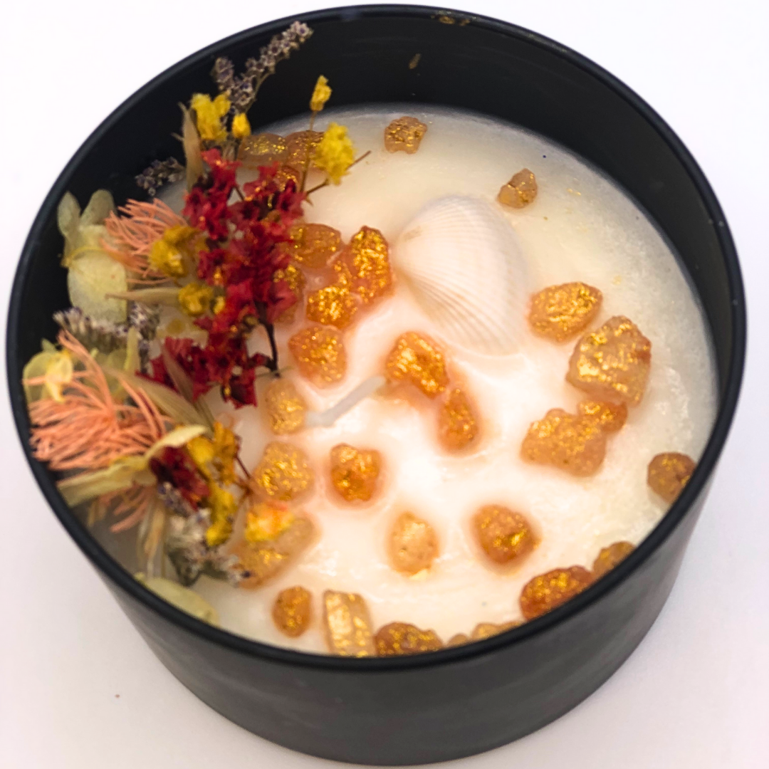 A double-wick white candle in a black jar, topped with dried yellow and red florals, small seashells, and golden rocks.