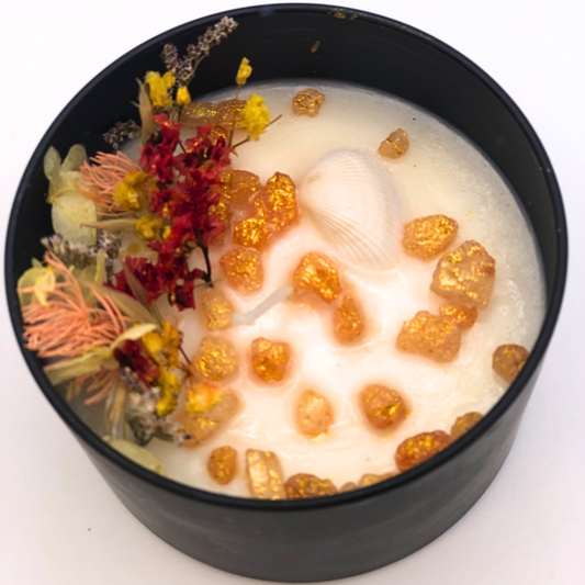 A double-wick white candle in a black jar, topped with dried yellow and red florals, small seashells, and golden rocks.