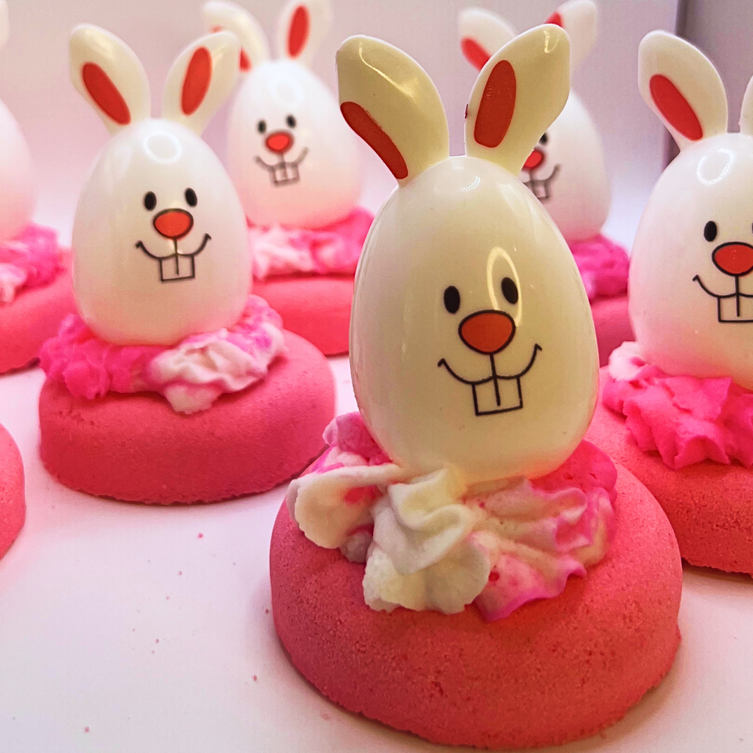 A pink donut-shaped bath bomb topped with pink and white bubble bath icing and a bunny-shaped egg decoration.