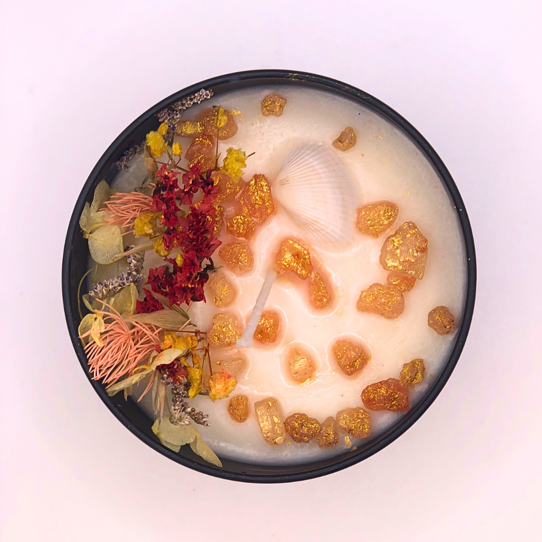 A double-wick white candle in a black jar, topped with dried yellow and red florals, small seashells, and golden rocks.