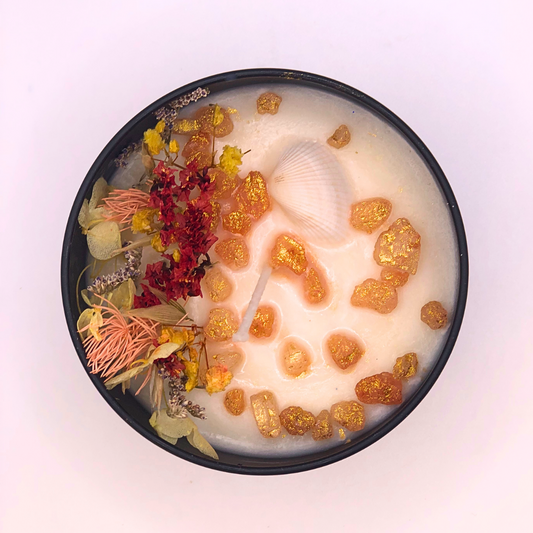 A double-wick white candle in a black jar, topped with dried yellow and red florals, small seashells, and golden rocks.