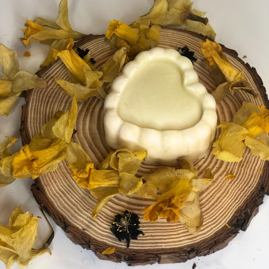 A white heart-shaped soap bar placed on a piece of wood, decorated with dried yellow flowers.