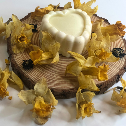 A white heart-shaped soap bar placed on a piece of wood, decorated with dried yellow flowers.